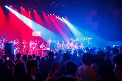 Het NSJO staat onder rode en blauwe lampen op het podium te spelen. Voor hen staan veel mensen te kijken.