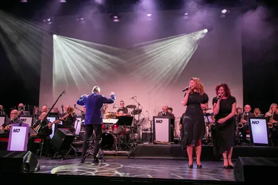 Twee zangeressen zingen op het podium voor een orkest, terwijl de dirigent in een blauw jasje het orkest dirigeert onder helder licht.