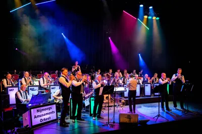 Een groot orkest speelt op een verlicht podium met koperblazers, toetsen en slagwerk, terwijl de dirigent het Nijmeegs Amusementsorkest aanstuurt.
