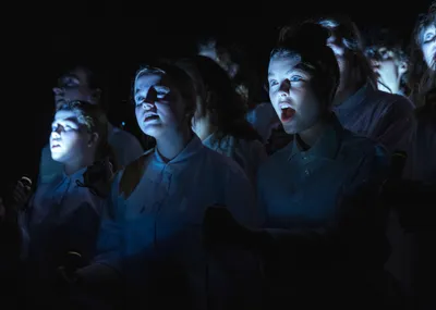 In een donkere setting staan jonge mensen in witte kleding. Ze kijken naar een publiek. Een meisje heeft haar mond open.