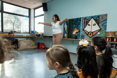 Een dansdocent laat kinderen in een klaslokaal vrolijk meebewegen tijdens een dansles met muziek en kleurige schilderijen aan de muur.