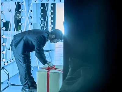A blue-lighted picture of a man unboxing a present, standing with his head against the wall.