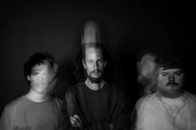 De drie mannen van de band kijken in de camera, door een effect is hun beweging nog wazig langs hen heen te zien.