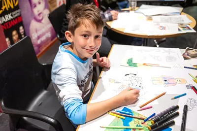 Een jongen zit aan een tafel vol kleurpotloden en tekenspullen en kijkt glimlachend naar de camera terwijl hij tekent