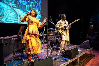 Een zangeres in een kleurrijke jurk en een gitarist treden op op een verlicht podium met een groot projectiescherm achter hen