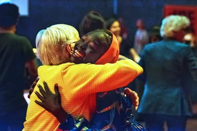 Twee mensen omhelzen elkaar stevig; één persoon lacht breed terwijl de achtergrond vervaagd is door beweging en licht