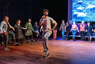Een groep volwassenen staat in een rij hand in hand terwijl een dansleider voor hen danst en aanwijzingen geeft op een houten podiumvloer