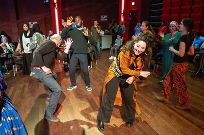 Drie mensen dansen enthousiast in een volle zaal, met andere bezoekers klappend en lachend op de achtergrond