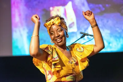 Een vrouw in een felgele, kleurrijke outfit danst op een podium. Ze glimlacht breed en heft haar armen in de lucht terwijl ze meebeweegt op de muziek.