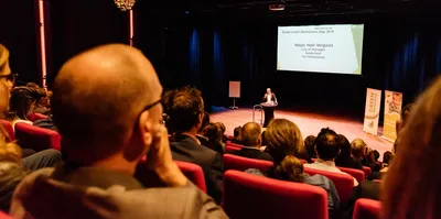 Een man in een grijs pak staat achter een katheder op het podium van de Lindenbergzaal en houdt een presentatie voor publiek