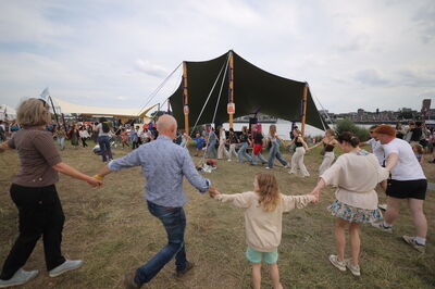 Grote groep mensen van alle leeftijden die hand in hand danst op een festivalterrein.