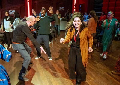 Een groep volwassenen danst lachend op een houten vloer in een zaal met rood licht, terwijl anderen toekijken