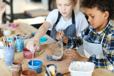 Twee kinderen schilderen met concentratie hun zelfgemaakte kleipot, met potjes verf en kwasten op tafel.