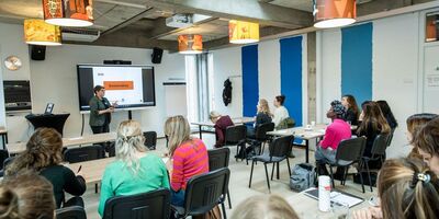 Een lichte vergaderruimte waarin iemand een presentatie houdt op een groot beeldscherm en mensen luisteren