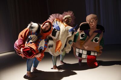 Dansers in kleurrijke, speelse kostuums met fantasiegezichten buigen naar voren op een zacht verlichte toneelvloer