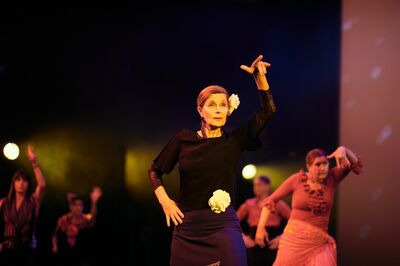 Vrouw danst expressief op podium, met andere dansers op de achtergrond.