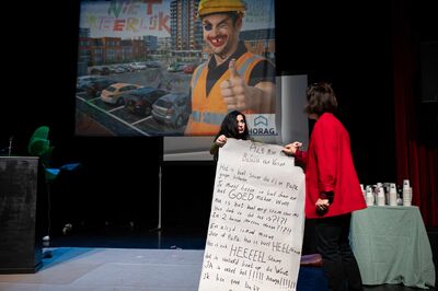 Acteur toont een groot handgeschreven vel papier aan een andere speler, met projectie van een bouwvakker op de achtergrond.