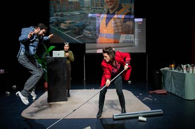 Twee acteurs in beweging op een podium met een grote projectie achter hen; een vrouw veegt met een trekker.