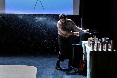Vrouw op toneel wordt natgespoten bij een tafel met koffiekannen en bekers tijdens een theatrale scène.