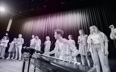 Een man zit achter een keyboard, achter hem staan mensen met kabelmicrofoons te zingen. 