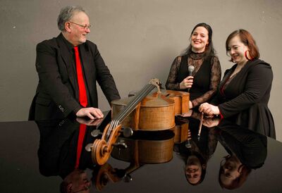 De drie muzikanten staan bij de piano en dragen zwarte kleren met rode accenten.