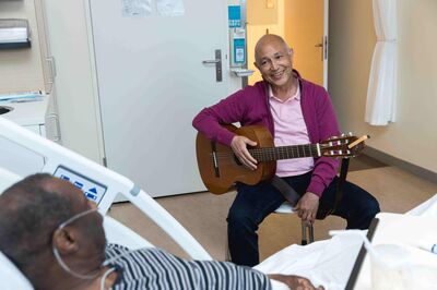 Een muzikant met zijn gitaar op schoot lacht naar een patiënt in een ziekenhuisbed.