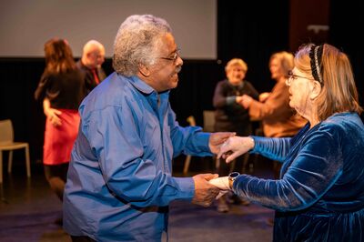 Een oudere man en vrouw dansen samen. Ze hebben elkaars handen vast en lachen. Op de achtergrond dansen meer paren.