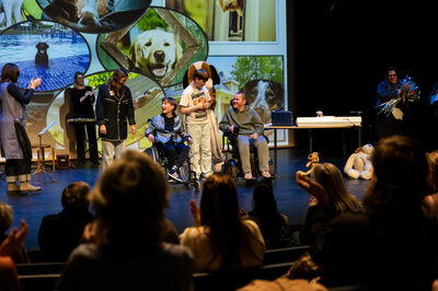 Jongeren in rolstoelen en begeleiders optreden op een podium met grote hondenfoto’s achter hen