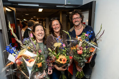 Drie mensen met grote bossen bloemen lachen na een presentatie of viering