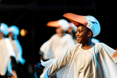 Kind danst in vogelkostuum met een grote oranje snavel, met anderen op de achtergrond