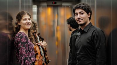 Een jonge vrouw en een jongen staan in de lift en kijken met een lieve lach richting de camera. 