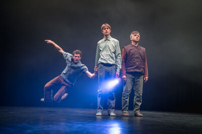 Twee mannen kijken emotieloos voor zich uit, terwijl een derde aan de hand van de ander vrolijk aan komt sliden.