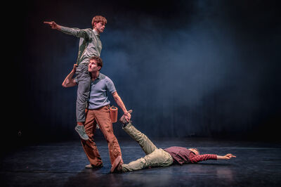 Drie mannen staan in een acrobatische houding, waarbij een man op de schouder van de ander zit. Daarbij wordt iemand anders op de grond aan zijn enkels voortgesleept.