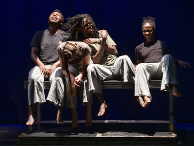 Vier dansers zitten op een bank en kijken intens in helder podiumlicht.