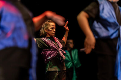Een kind op een podium danst geconcentreerd, gekleed in een paars kostuum met felgekleurde schmink op het gezicht