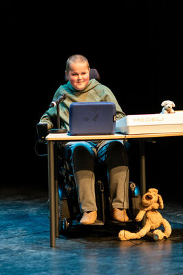 Jongere in een rolstoel zit achter een tafel op een podium met laptop, microfoon en knuffelhond