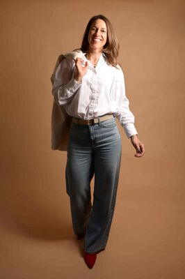 Een vrouw heeft haar jas om haar schouder gehangen en kijkt lachend en vol zelfvertrouwen de camera in.