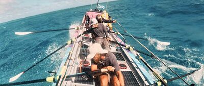 Drie mannen roeien op een oceaanroeiboot in een woeste zee, met opspattend water over het dek.
