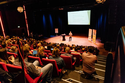 Beeld van het zijaanzicht van het podium van de Lindenbergzaal waar een pan in een grijs pak een presentatie geeft.