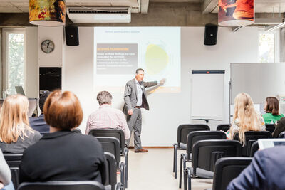 Een man in een grijs pak geeft een presentatie voor een groep mensen die op stoelen zitten. De ruimte is licht. 