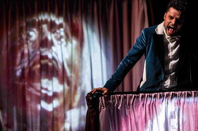 Een acteur in een blauw colbert schreeuwt expressief boven een theaterdoek, terwijl een groot, angstig gezicht op de achtergrond wordt geprojecteerd.