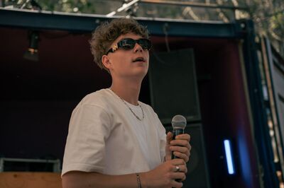 Een jonge man in een wit shirt met een zonnebril op heeft een microfoon in zijn handen. Hij staat op een podium.