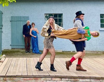 Twee acteurs dragen een liggende speler over een houten podium, terwijl andere spelers verbaasd toekijken bij een theatrale scène.