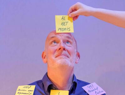 Een man kijkt naar boven, naar een memoblaadje met "al het moois" erop geschreven, dat door een hand boven zijn hoofd gehouden wordt.