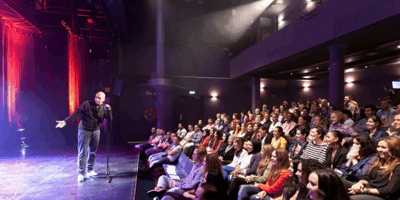 Publiek zit op de tribune in de Steigerzaal en kijkt naar een man met microfoon op het podium die in theaterlicht staat.