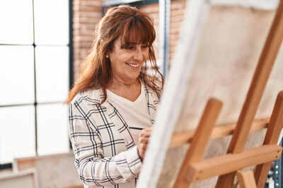 Een vrouw schildert met een glimlach op een doek. De doek staat op een schildersezel in een lichte atelierruimte.