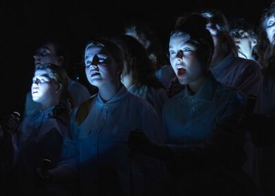 In een donkere setting staan jonge mensen in witte kleding. Ze kijken naar een publiek. Een meisje heeft haar mond open.