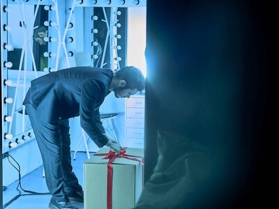 A blue-lighted picture of a man unboxing a present, standing with his head against the wall.