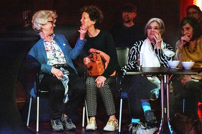 Vier oudere vrouwen zitten samen aan een tafel en kijken met vrolijke en nieuwsgierige gezichten naar iets buiten beeld