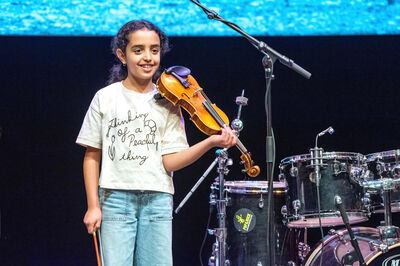 Een meisje op een podium houdt glimlachend een viool vast, met drums en microfoons achter haar